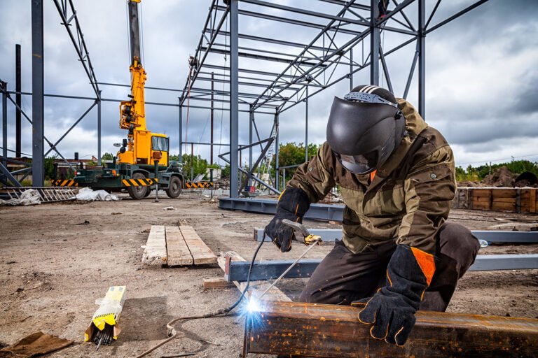 A young man welder in brown uniform, welding mask and welders leathers, weld metal with a arc welding machine at the construction site, blue sparks fly to the sides